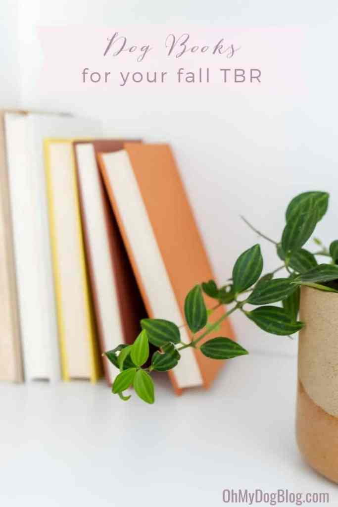 A stack of books leans against the edge of a bookshelf slightly out of frame. The spines are fall colors: yellow, tan, brown, and orange. In the foreground, a vine drapes out of a tan and gold pot. The text overlay reads: Dog books to add to your fall TBR.